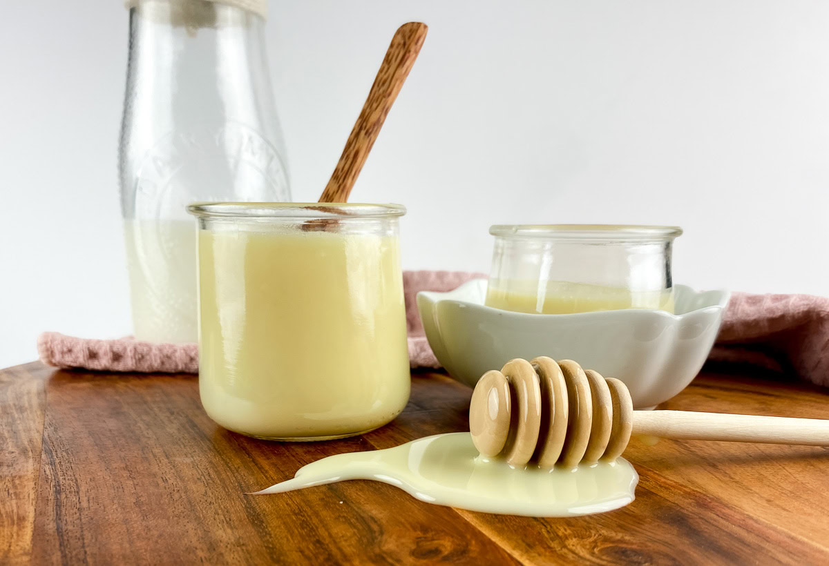 Homemade sweetened condensed milk in glass jars on a wooden surface, with a honey dipper and milk bottle in the background.
