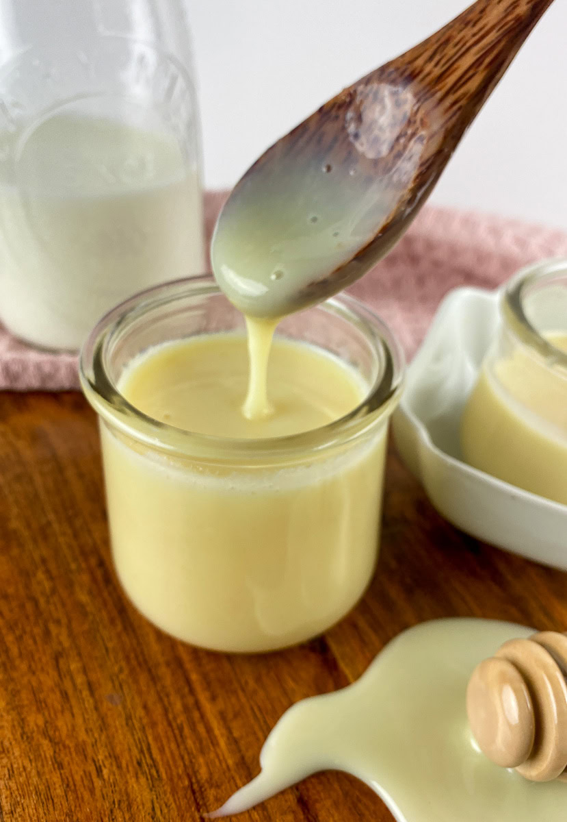 A wooden spoon drizzles creamy homemade sweetened condensed milk into a small glass jar, with another jar and a milk bottle in the background on a wooden surface.