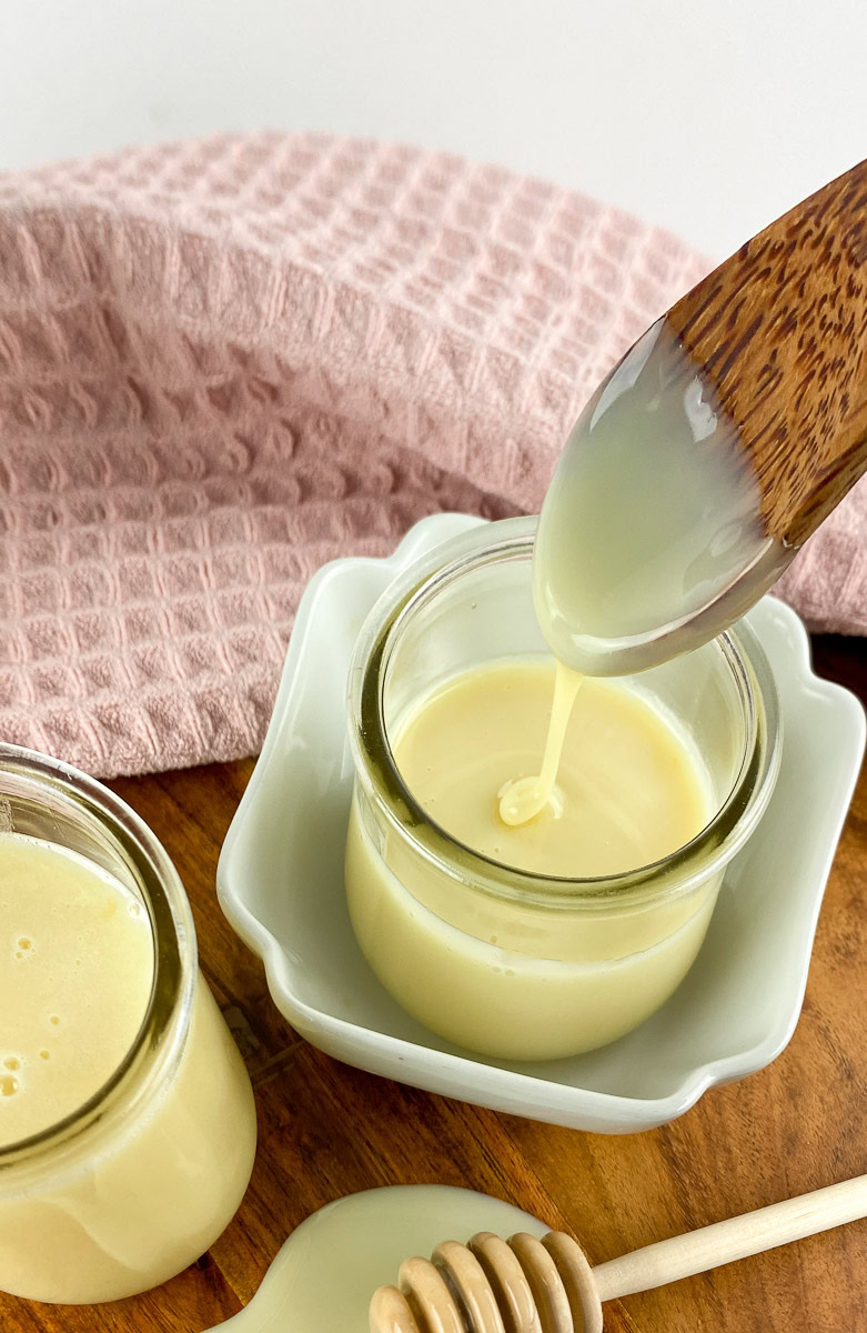 Homemade sweetened condensed milk in a glass jar with a wooden spoon drizzling the creamy milk, placed on a wooden board with a pink cloth in the background.