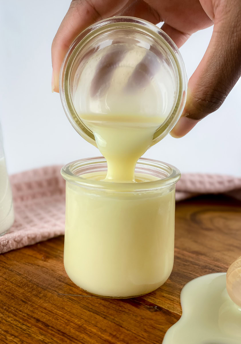 A close-up of a hand pouring thick, creamy homemade sweetened condensed milk from one jar into another on a wooden surface.