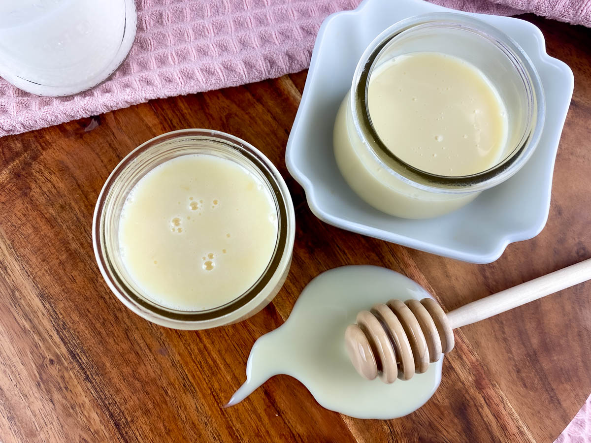 Top view of homemade sweetened condensed milk in glass jars with a honey dipper on a wooden board.