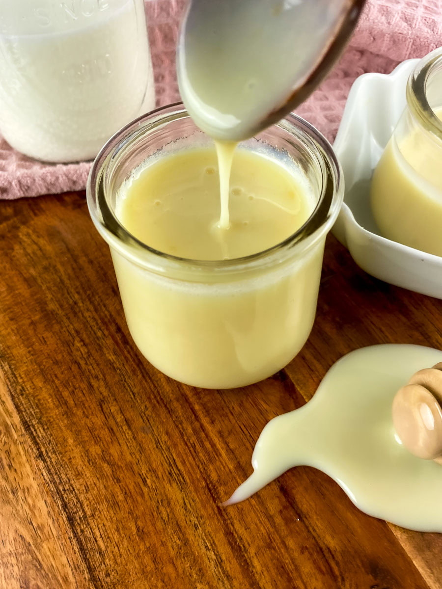 Pouring homemade sweetened condensed milk from a spoon into a glass jar on a wooden table, with a pink cloth and other jars in the background.