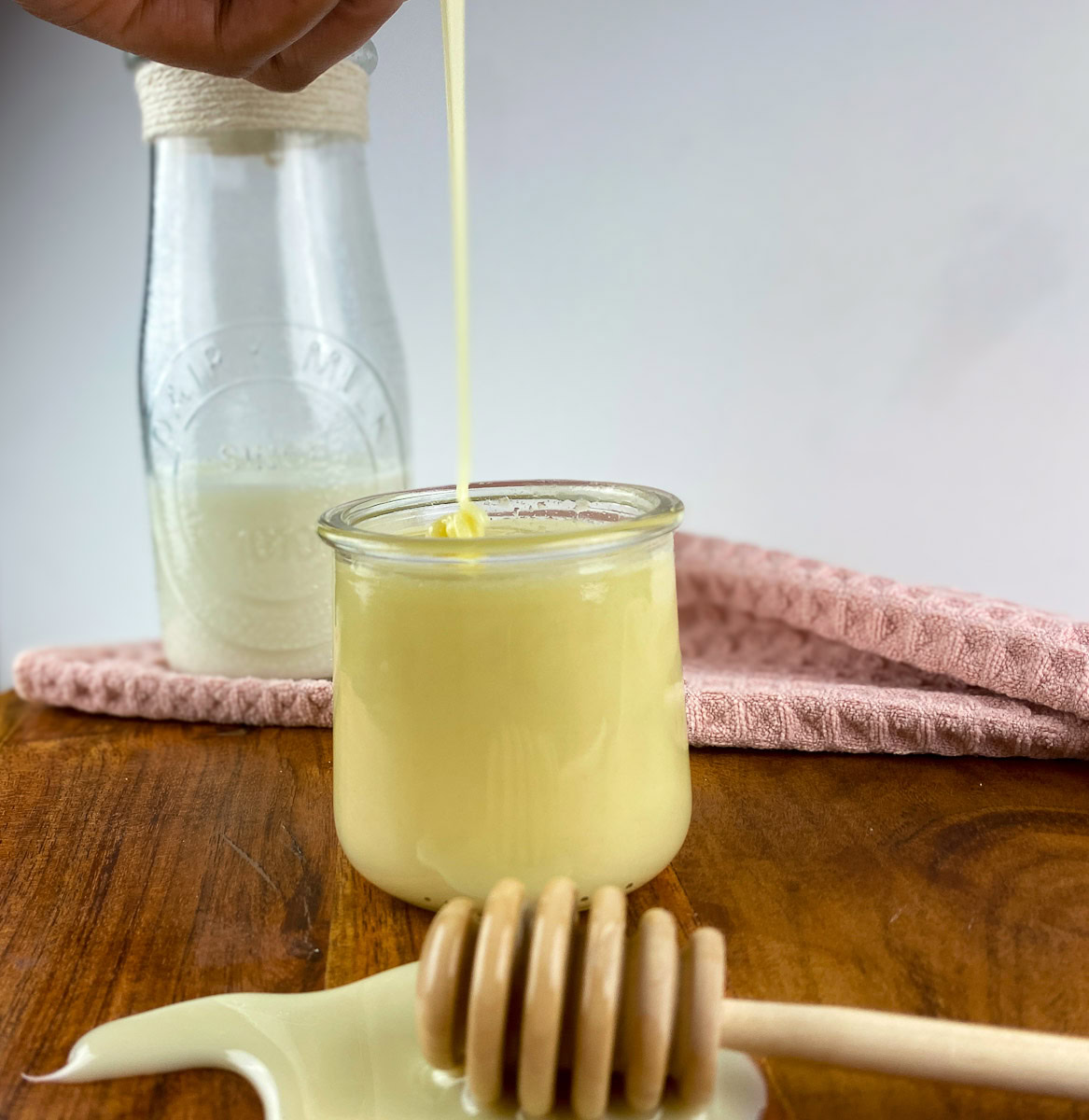 Thick stream of homemade condensed milk being poured into a glass jar on a wooden table, with a honey dipper and milk bottle in the background.